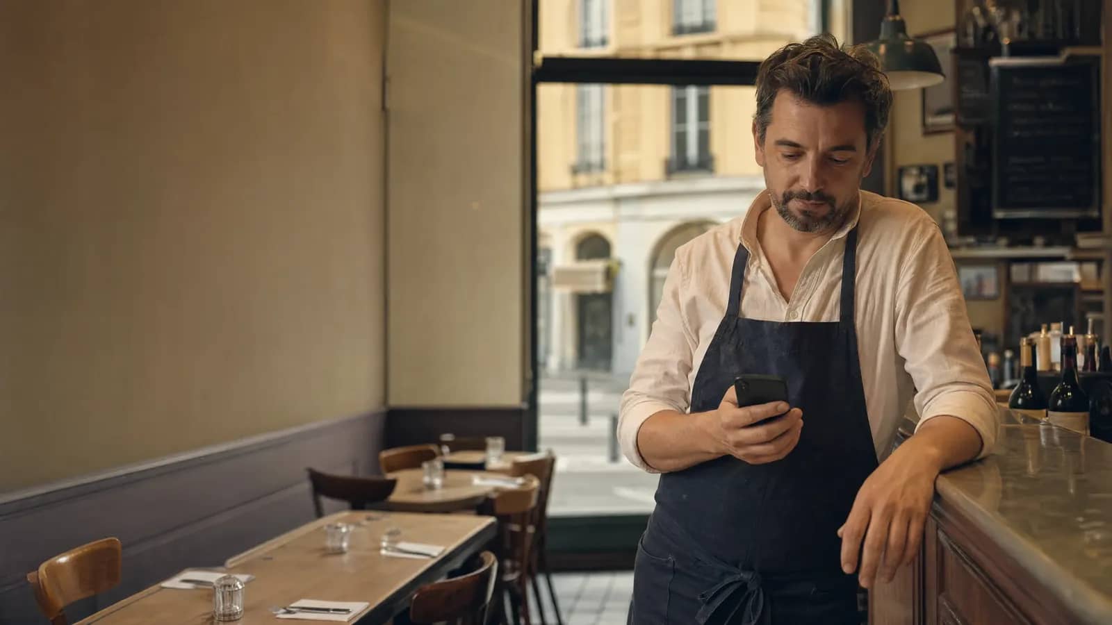 Restaurateur lyonnais au comptoir de son bistrot consultant son smartphone, façades de la Presqu'île par la fenêtre