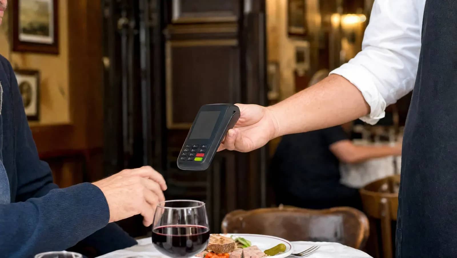 Serveur tendant un terminal de paiement à un client attablé dans un bistrot lyonnais, avec un verre de vin rouge et une assiette de cochonnaille.