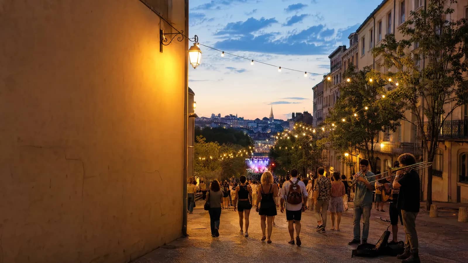 Foule en déambulation dans une rue lyonnaise illuminée de guirlandes au crépuscule pendant la Fête de la Musique, scène de concert en fond