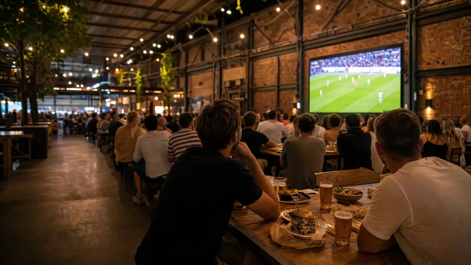 Foule de supporters attablés dans un food court lyonnais regardant un match de la Coupe du Monde 2026 sur écran géant, burgers et bières au premier plan