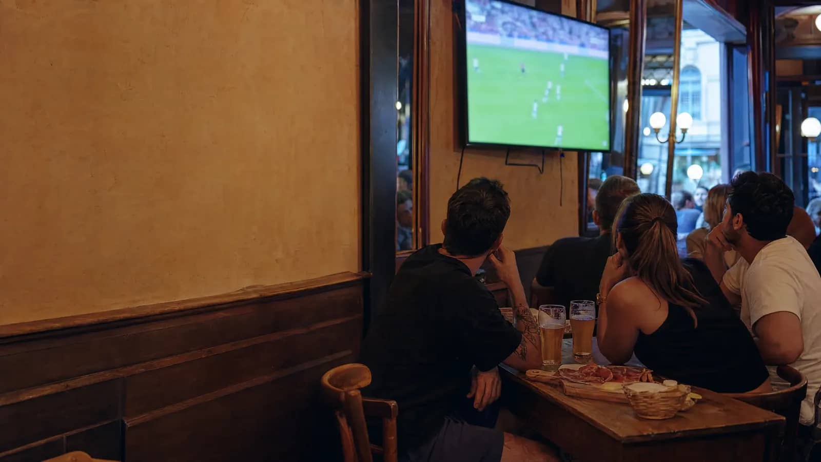 Groupe d'amis attablés dans une brasserie lyonnaise un soir d'été, regardant un match de la Coupe du Monde sur un écran, bières et planche de charcuterie sur la table.