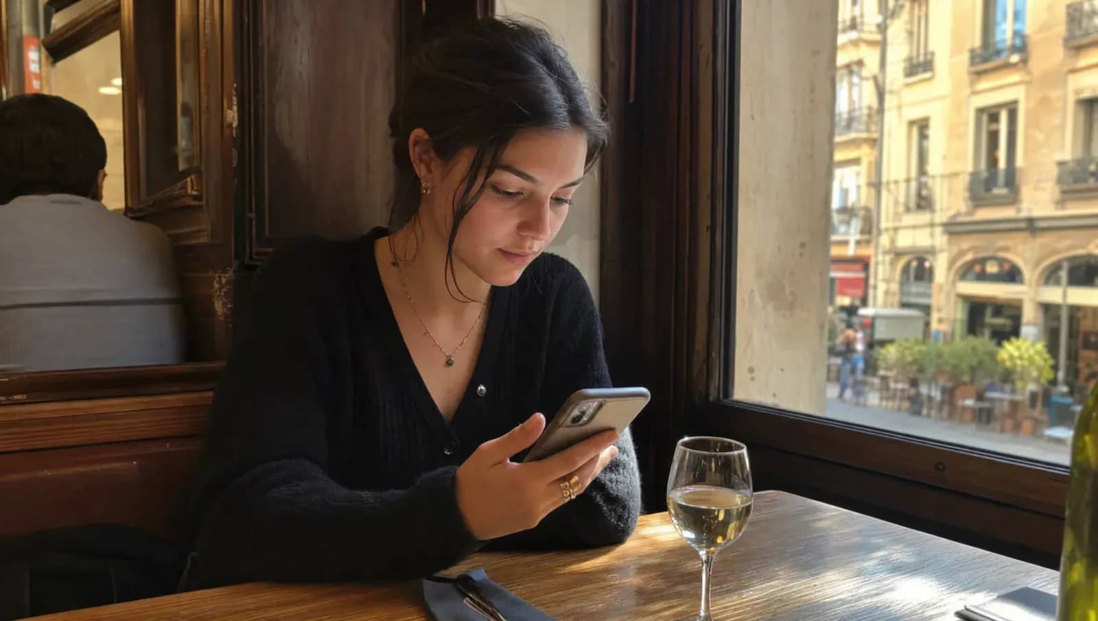 Cliente attablée dans un bistrot lyonnais cherchant un restaurant sur son smartphone, façades ocres par la fenêtre