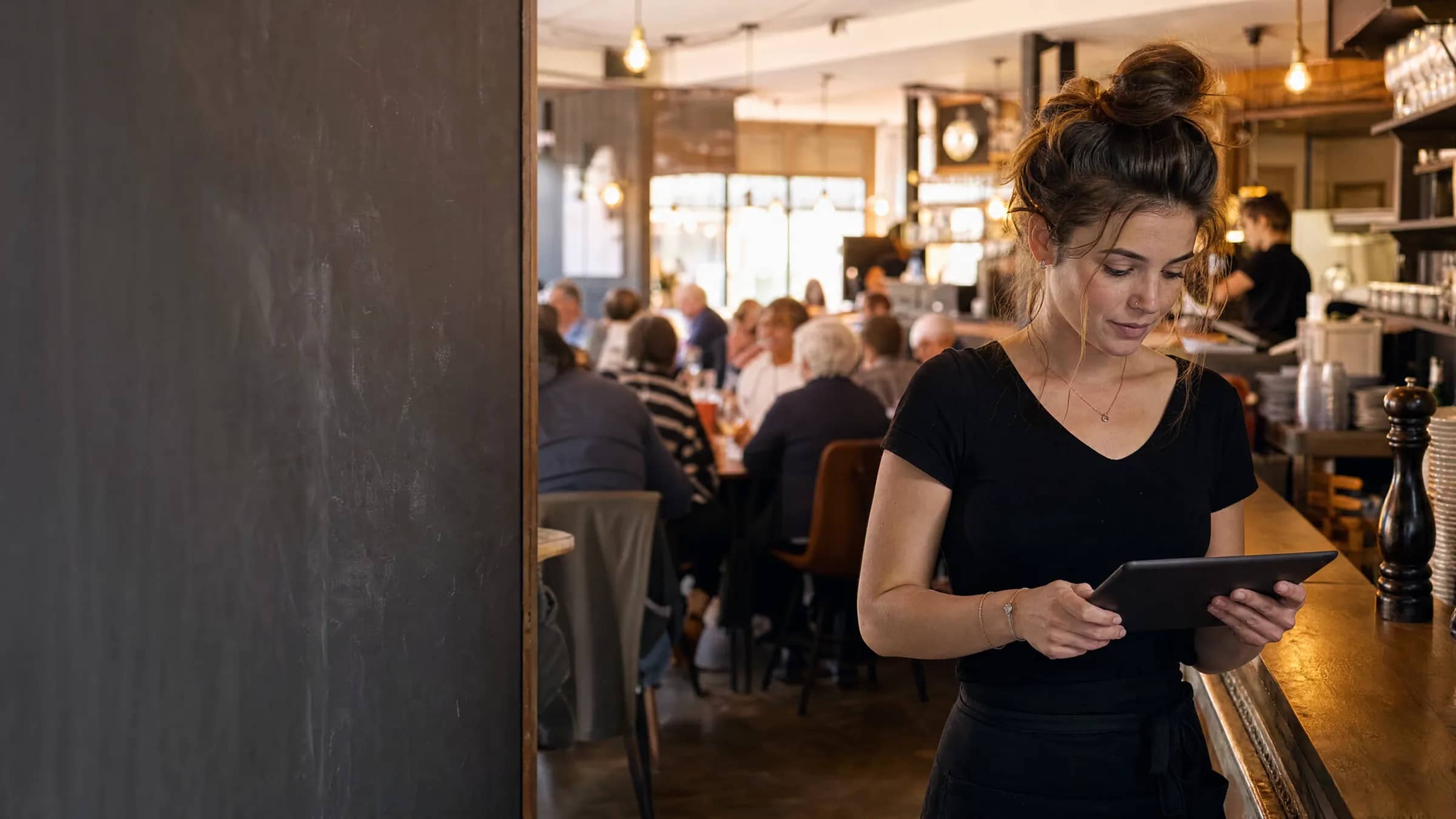 Une jeune restauratrice lyonnaise consulte une tablette derrière son comptoir pendant que sa salle se remplit, illustrant TheGoodSeat comme alternative à Restomatik qui génère vraiment de la demande.