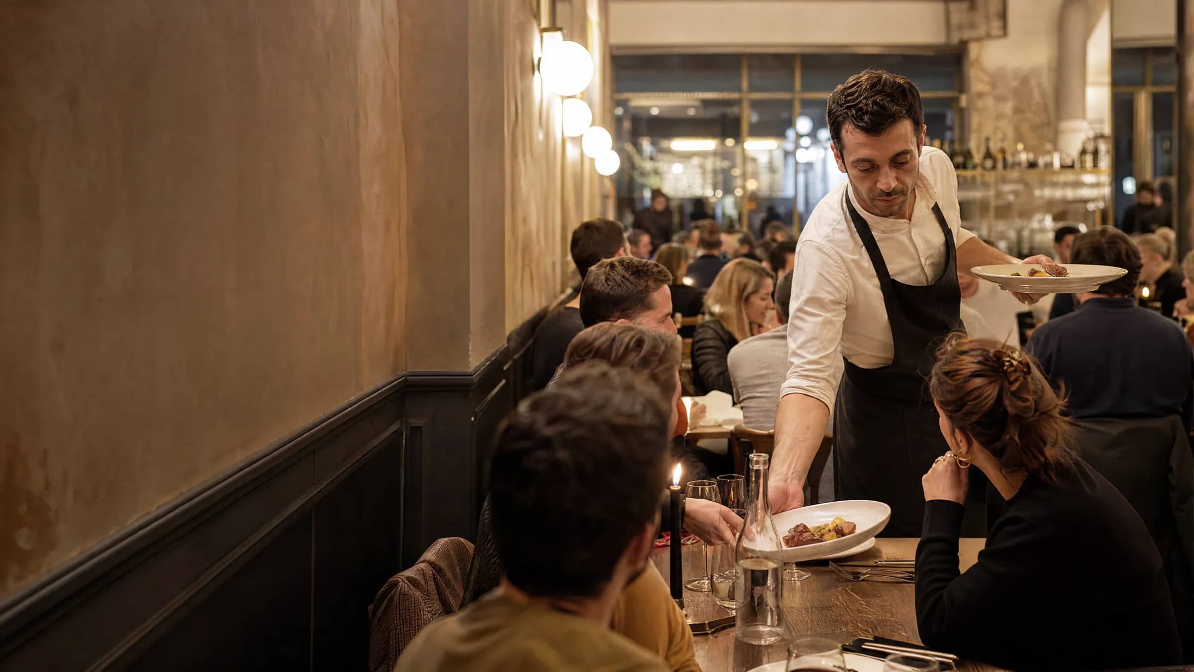 Un serveur apporte des assiettes à une table de dîneurs dans une salle de bistrot lyonnais bien remplie en service du soir, illustrant le remplissage des couverts par TheGoodSeat à côté de l'anti-gaspi de Too Good To Go