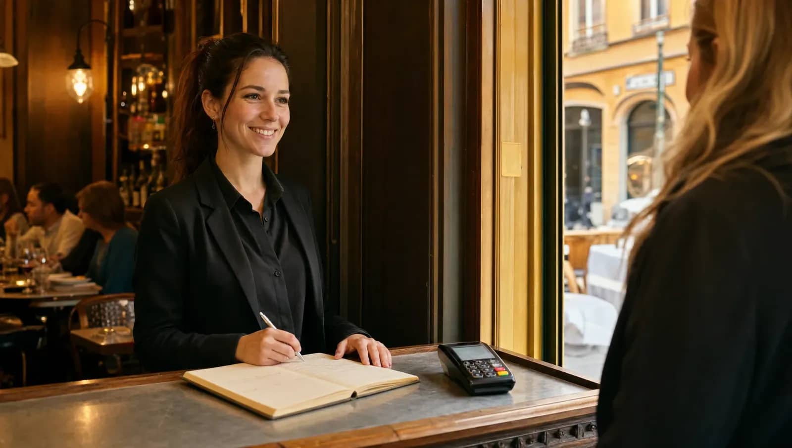 Hôtesse d'un bistrot lyonnais prenant une réservation, carnet ouvert et terminal de paiement sur le comptoir