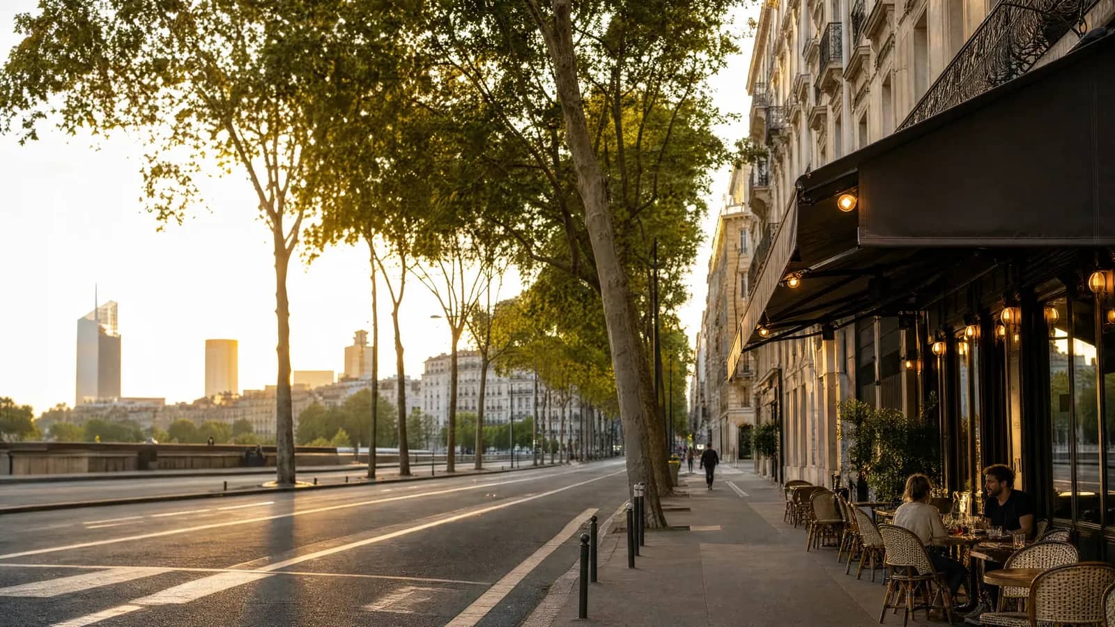 Terrasse de bistrot sur un boulevard arboré du 3e arrondissement de Lyon, avec les tours de la Part-Dieu au coucher du soleil