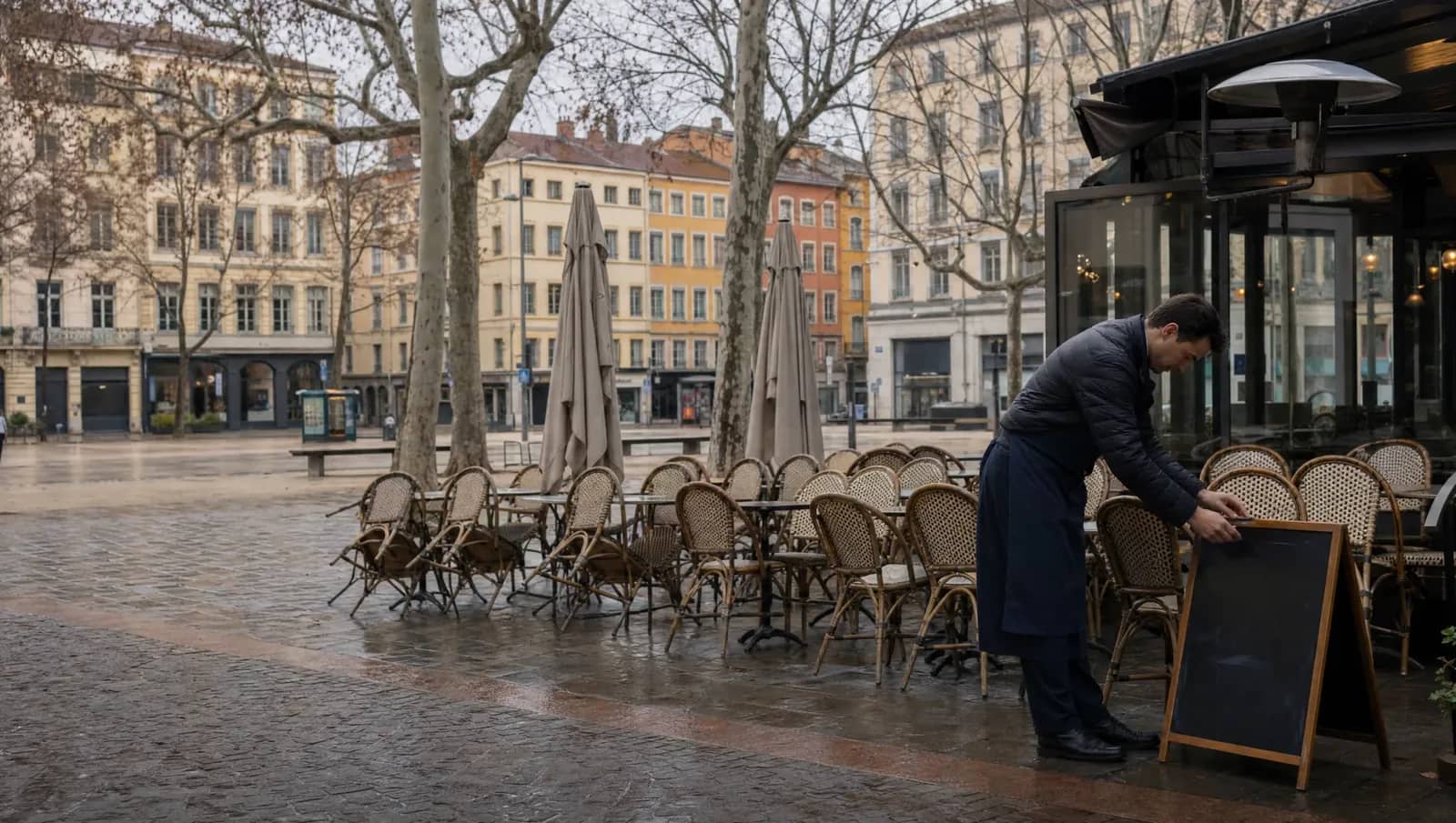 Terrasse de café déserte sur une place de la Presqu'île lyonnaise un matin gris de janvier, un serveur installe une ardoise.
